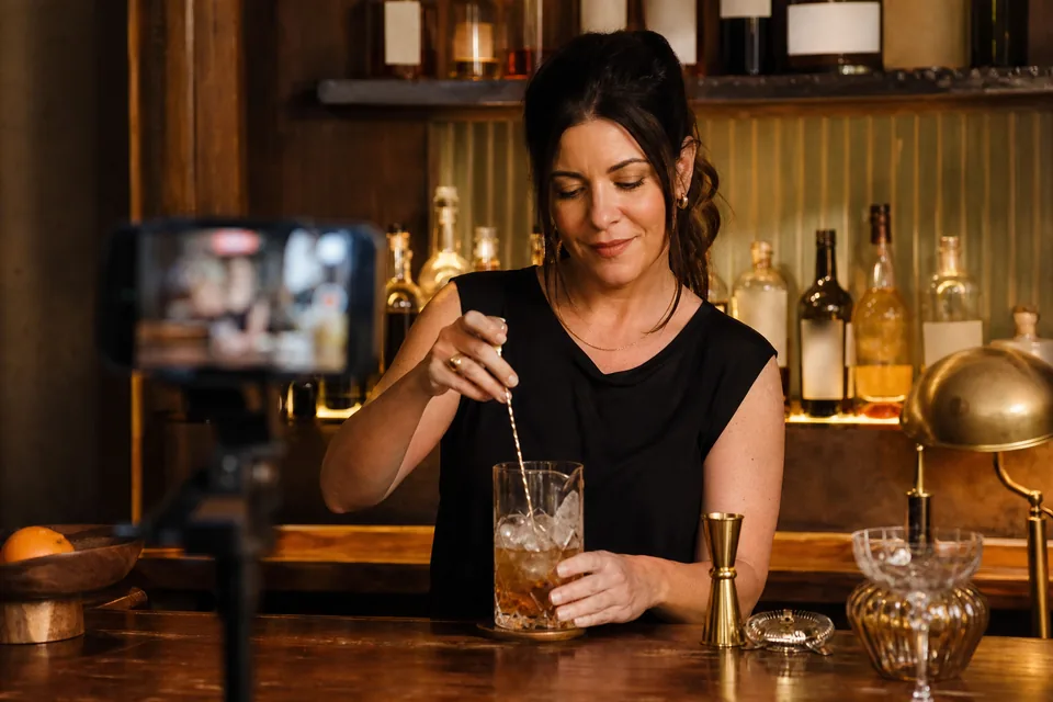 Bartender creator filming a cocktail recipe video on a phone tripod behind a home bar