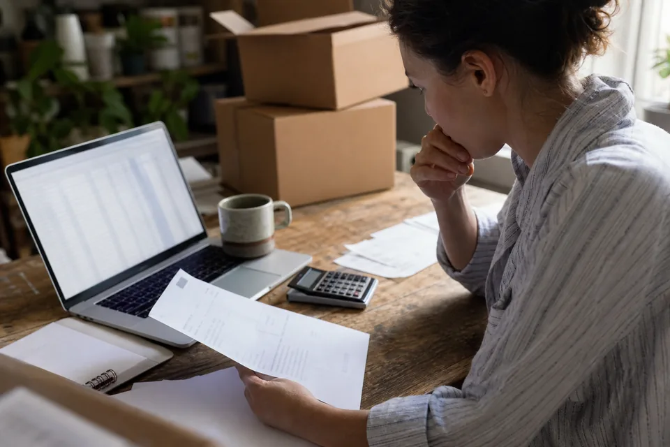 Small brand owner at a home office desk comparing monthly software costs on a laptop next to a calculator and shipping boxes